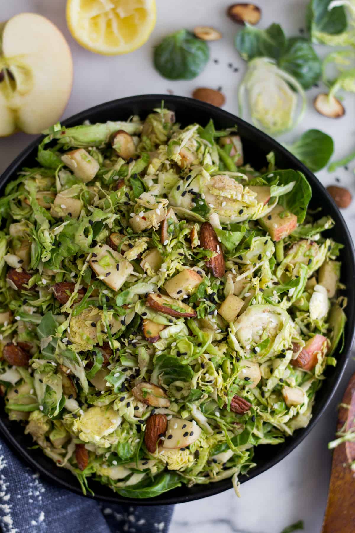 Big bowl with apple & shaved brussels sprouts salad surrounded by half an apple, half a lemon, brussels sprouts leaves, and almonds