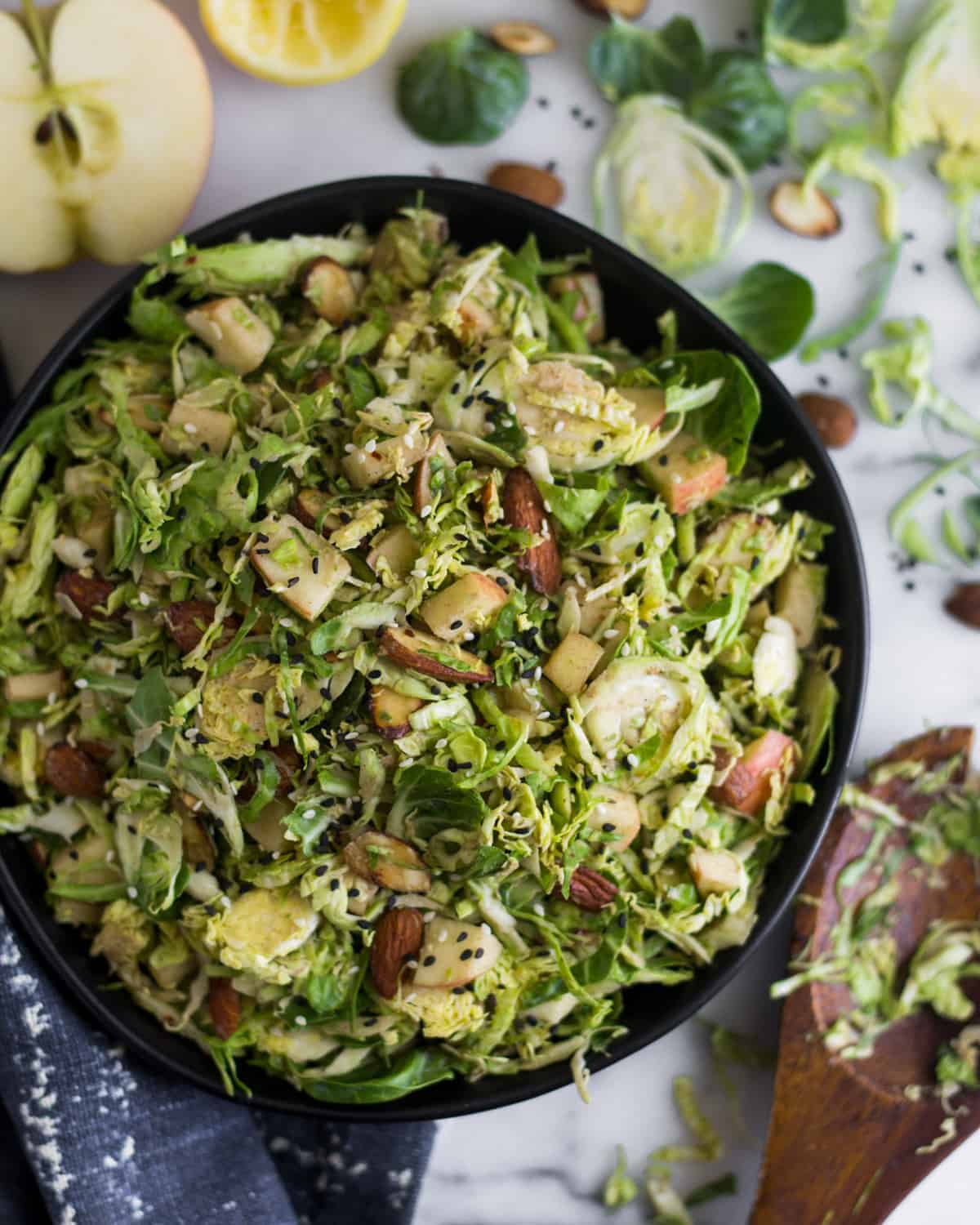 Big bowl with apple & shaved brussels sprouts salad surrounded by half an apple, half a lemon, brussels sprouts leaves, and almonds