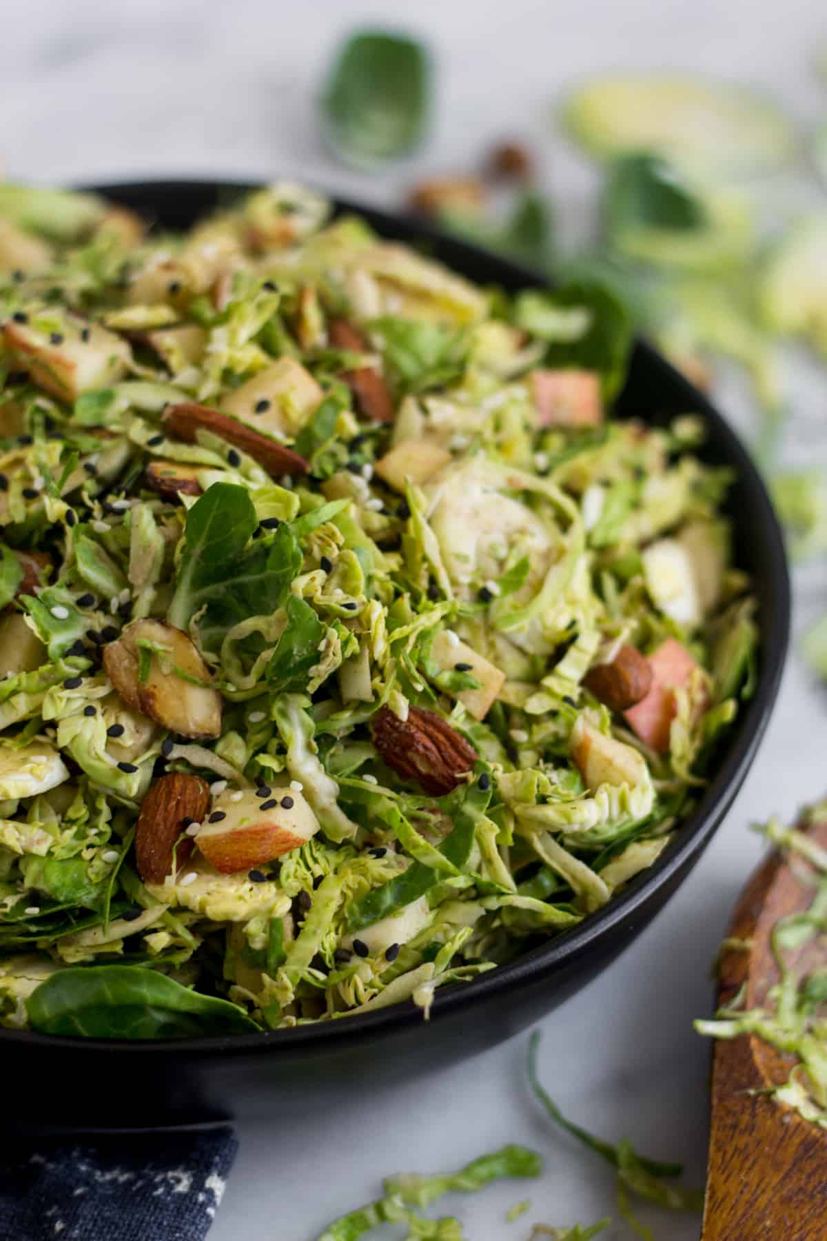 Close up of a black bowl with apple & shaved brussels sprouts salad with a wooden salad tong next to it