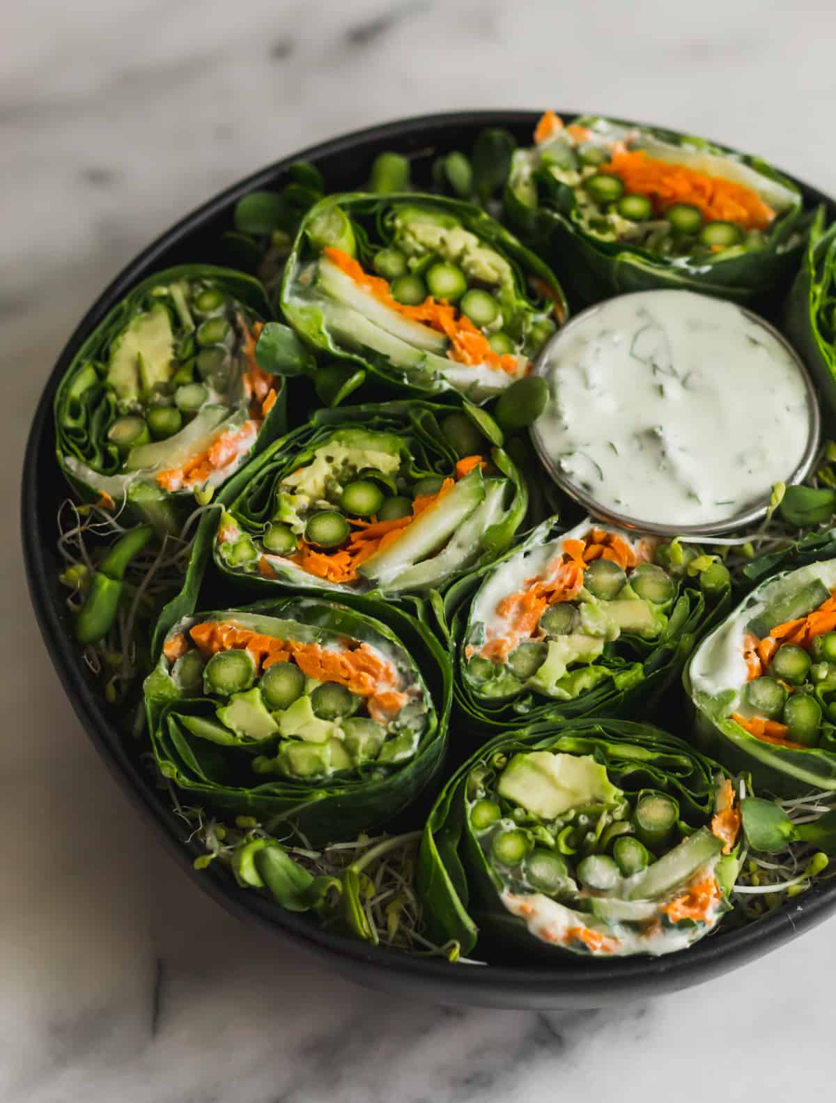Asparagus collard wraps in black bowl with a small cup of whipped feta on white marble