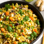 Bowl of broccoli fried rice on a white linen with chopsticks next to it and behind it a stalk of ginger, half a lime, green onions, and bowl of coconut aminos.