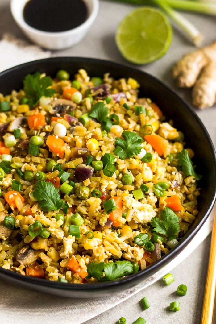 Bowl of broccoli fried rice on a white linen with chopsticks next to it and behind it a stalk of ginger, half a lime, green onions, and bowl of coconut aminos.