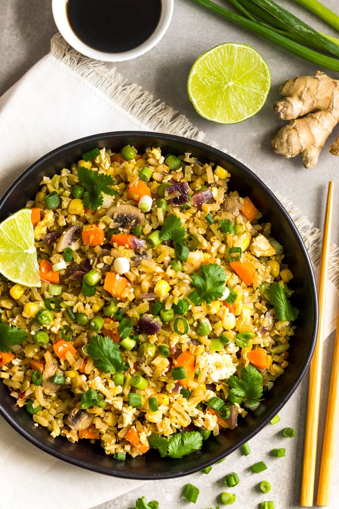 Overhead shot of a large black bowl filled with veggies fried rice topped with cilantro and a lime wedge. Around it is chopsticks, half a lime, ginger, bowl of coconut aminos, and green onions.