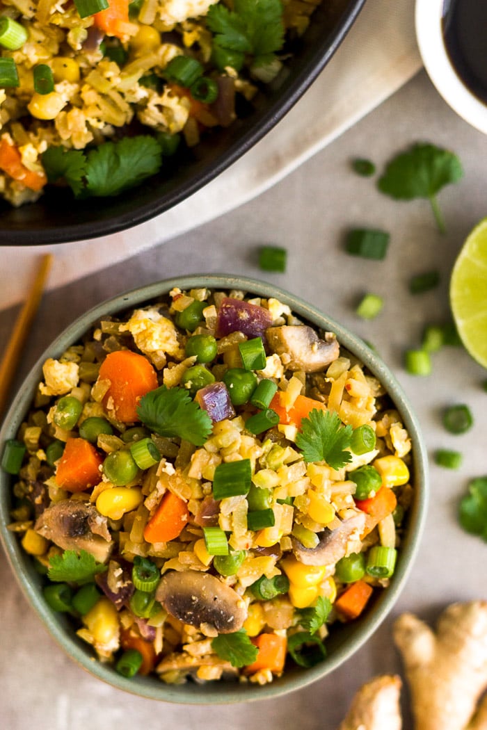 Small bowl of vegetable fried rice next to a large bowl of it. It is surrounded by ginger, green onions, lime juice, and coconut aminos.