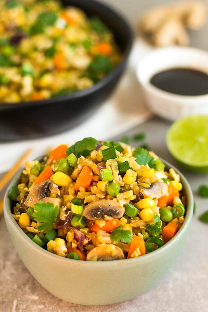 Small green bowl of broccoli fried rice topped with cilantro. Behind it is a large bowl of rice, half a lime, bowl of coconut aminos, and fresh ginger.