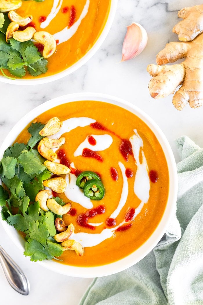 Overhead of two bowls of curried butternut squash soup topped with cilantro, cashews, coconut milk, hot sauce, and jalapeños. Next to it is a green towel, fresh ginger, and a clove of garlic.