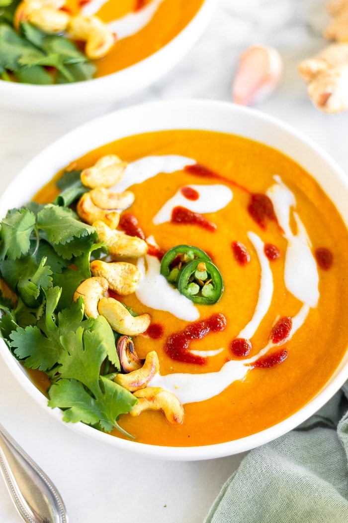 Bowl of curried butternut squash soup topped with coconut milk, hot sauce, sliced jalapenos, cashews, and cilantro. Behind it is another bowl and a clove of garlic and fresh ginger.