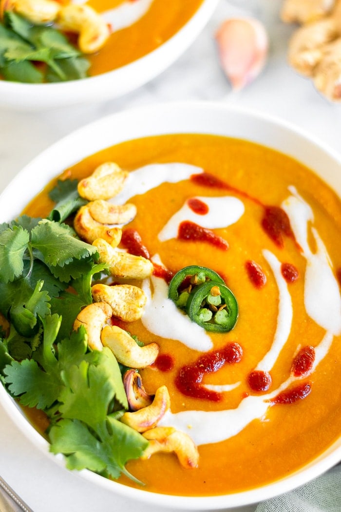 A close up of a bowl of coconut curry butternut squash soup topped with cilantro, whole cashews, coconut milk, hot sauce, and jalapenos. Behind it is another bowl, garlic, and ginger.