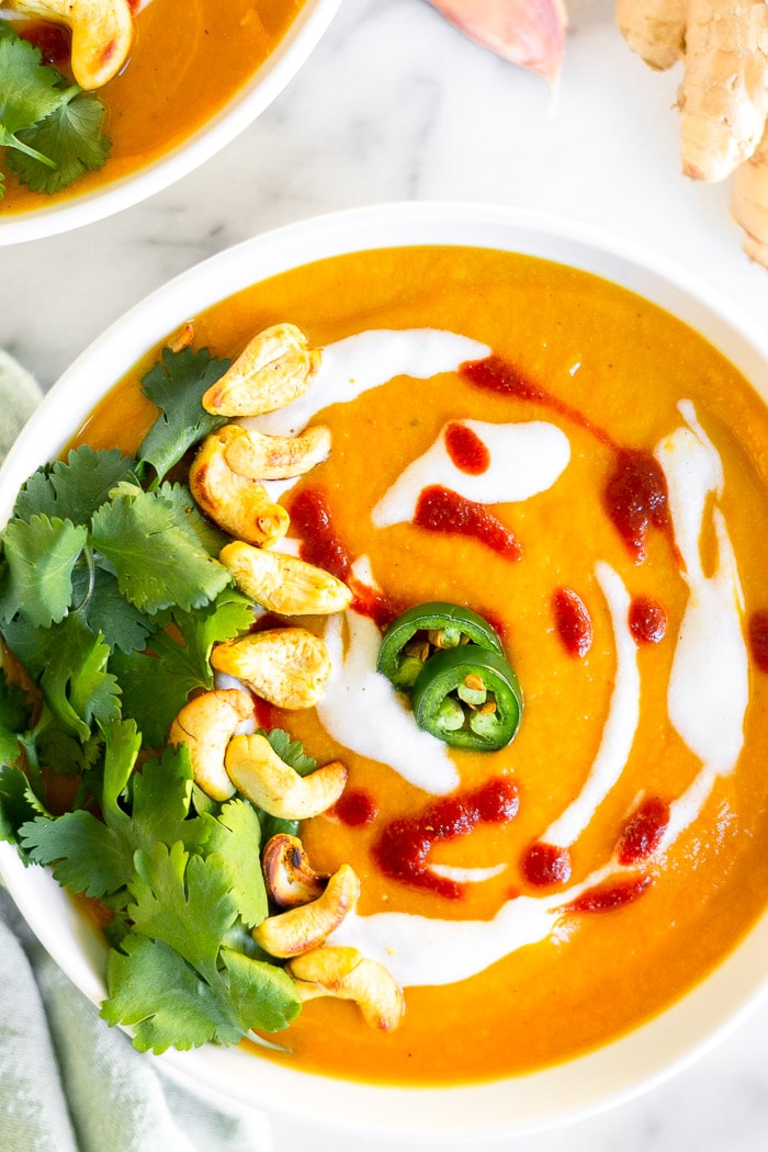 Coconut curry cauliflower soup topped with coconut milk, hot sauce, cilantro, cashews, and jalapenos. Behind it is another bowl of soup, fresh ginger, and a clove of garlic.