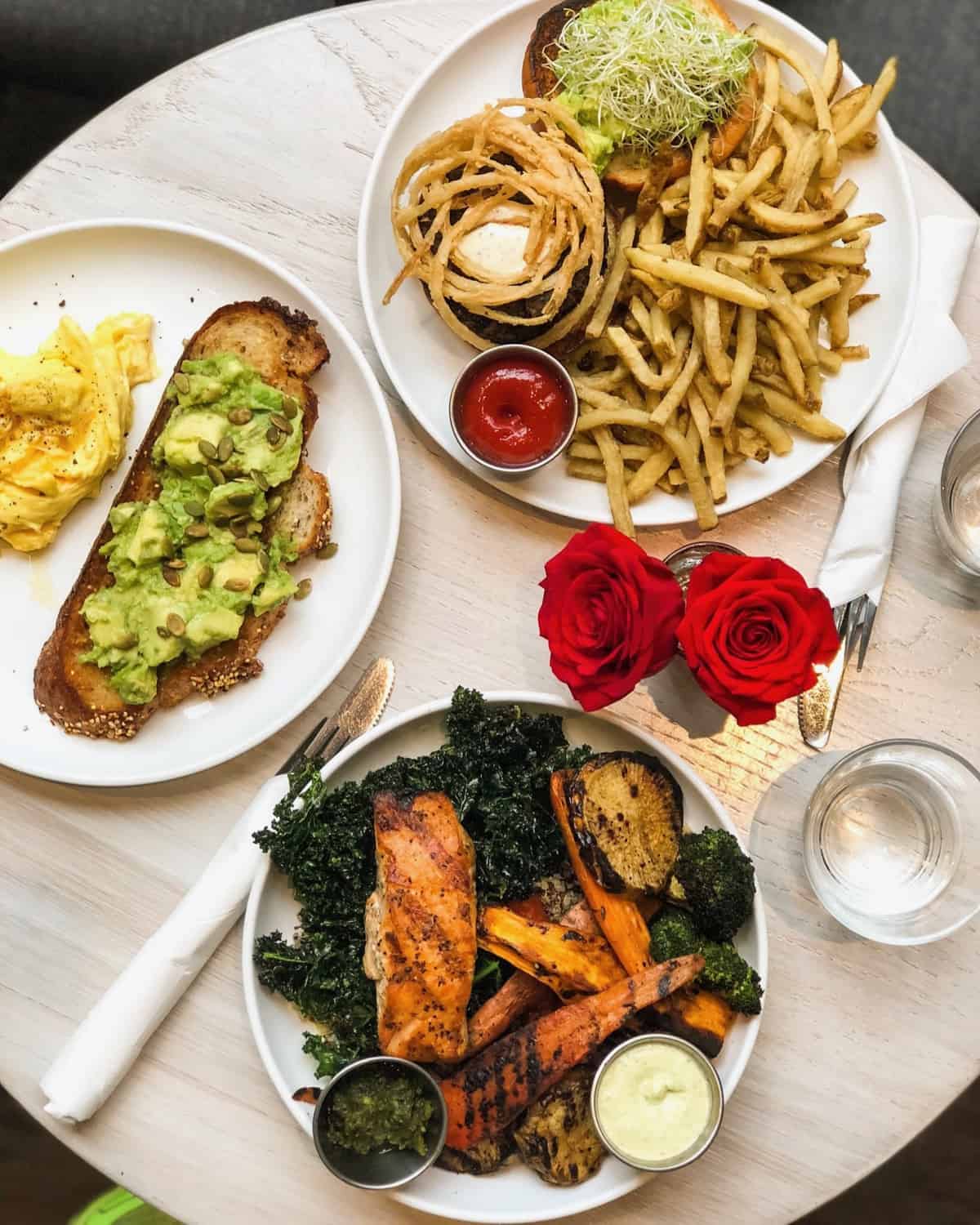 Table of a big bowl of veggies with grilled salmon on it, avocado toast with scrambled eggs, and a veggie burger with french fries