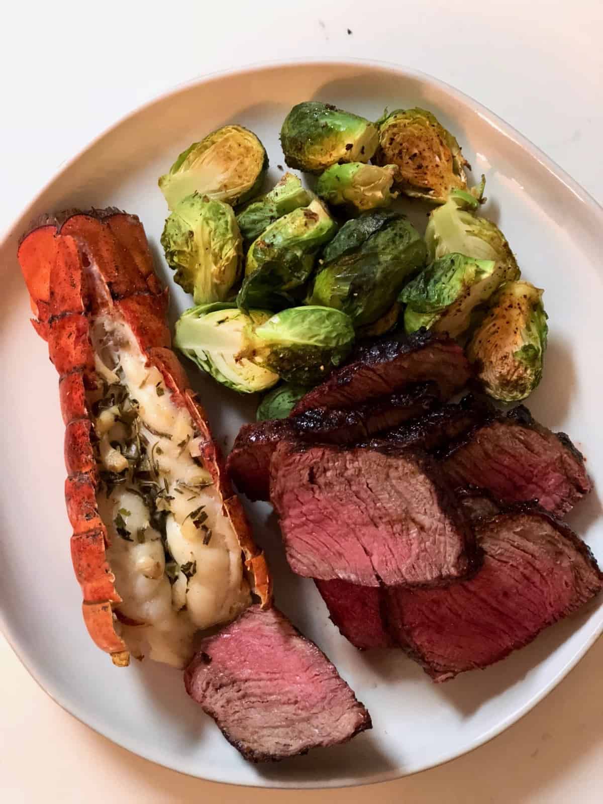 White plate of steak, brussel sprouts, and lobster