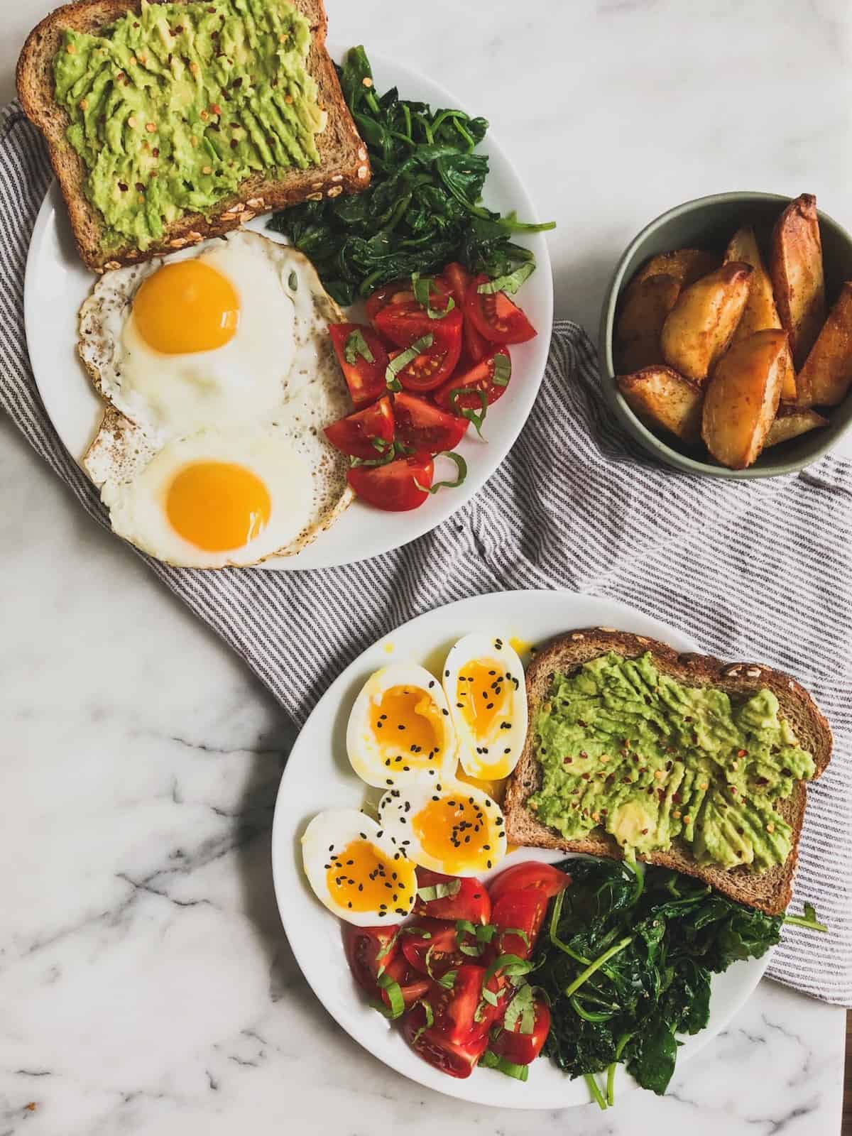 Two white plates with eggs, avocado toast, greens, and tomatoes with a small bowl of potato wedges on the side