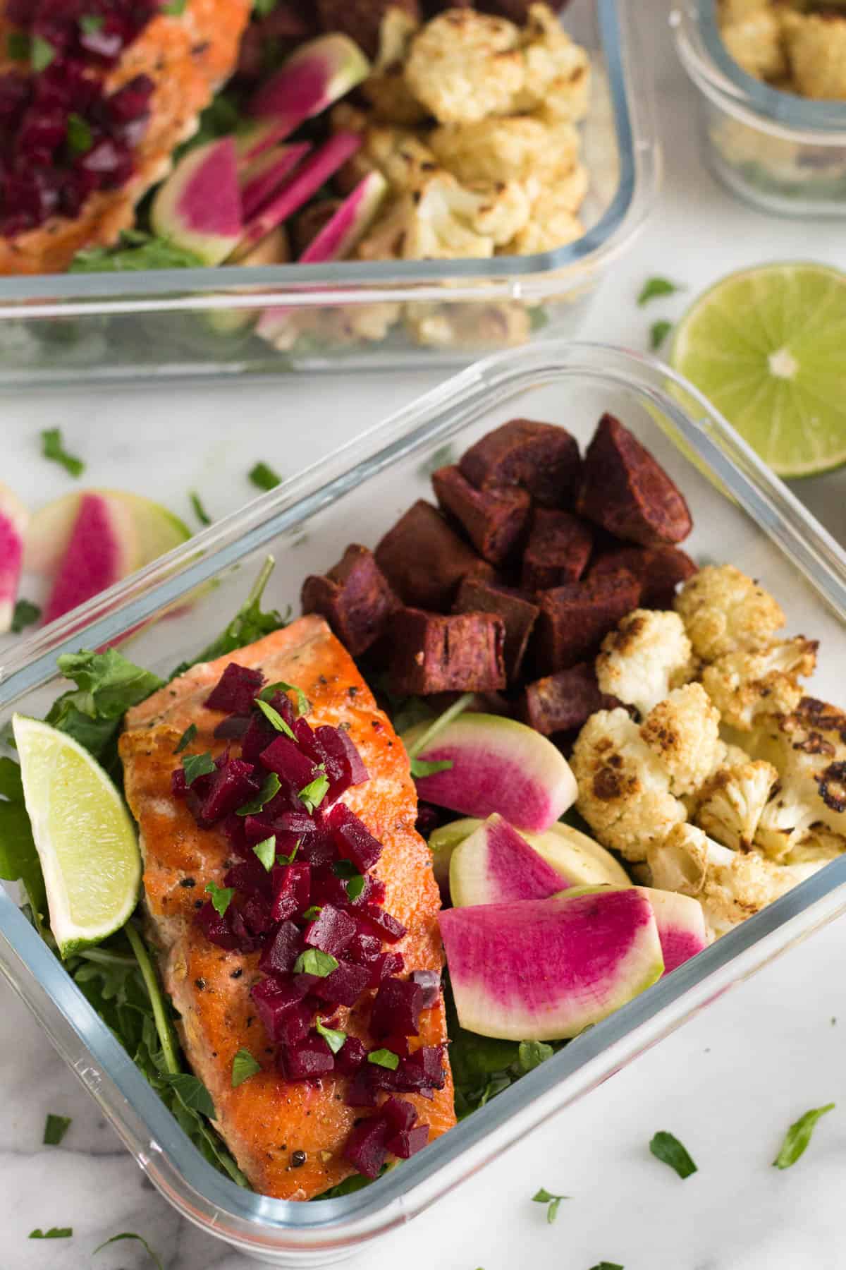 Two meal prep containers of greens, roasted veggies, and pan friend salmon topped with beets and a lime wedge.