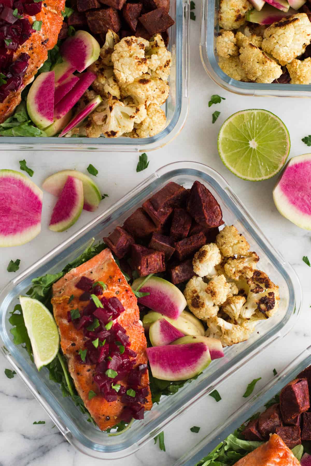 Multiple glass meal prep container filled with salmon and veggies on a white counter with limes and radishes on it
