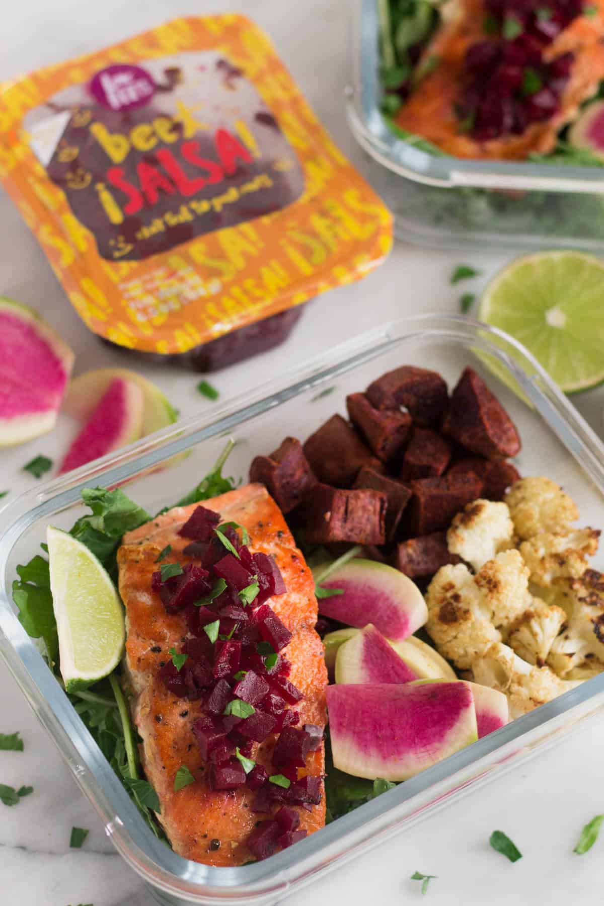 Glass container filled with greens, roasted veggies, and pan fried salmon with another container in the back and a package of precooked beets