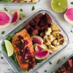 Glass meal prep container filled with salmon and veggies on a white counter with limes and radishes on it
