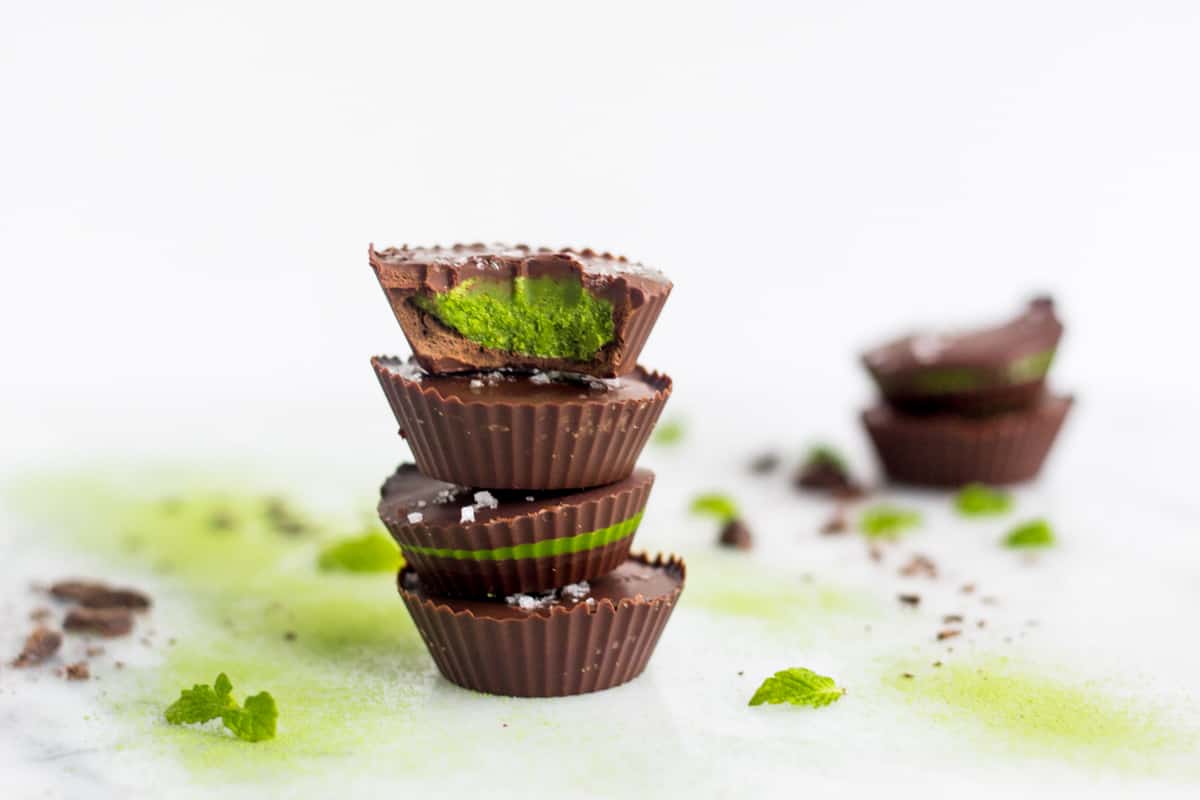 A stack of 4 Collagen Mint Matcha Coconut Butter Cups surrounded with chocolate and mint leaves with 2 in the background