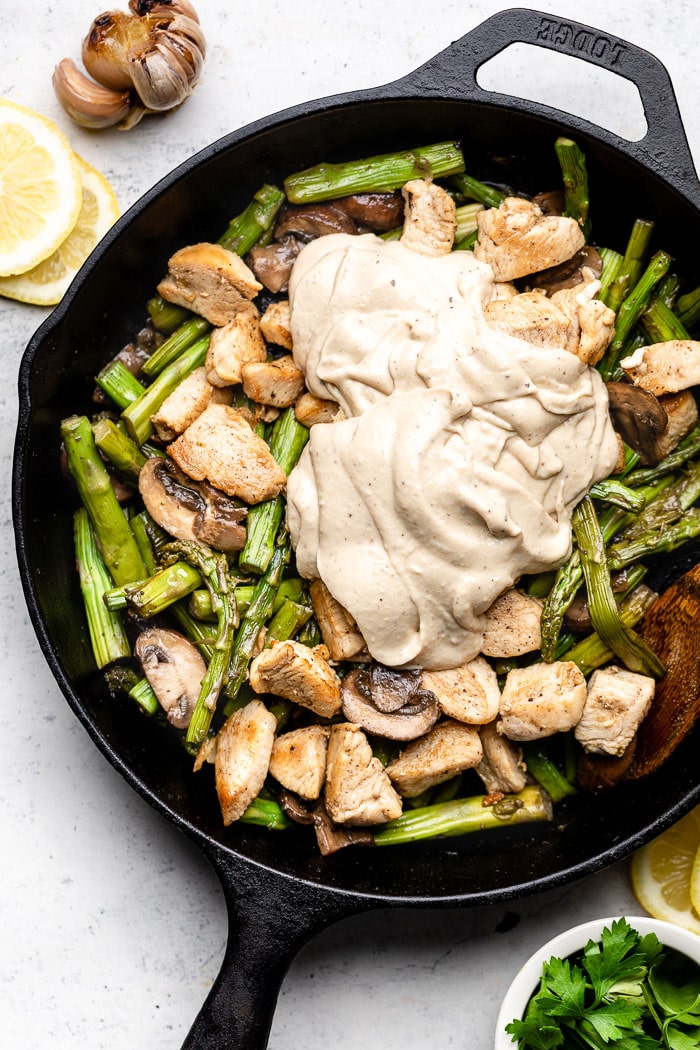 Chicken asparagus and mushroom in a large skillet topped with creamy lemon garlic sauce. Next to it is a bowl of parsley, sliced lemons, and a head of roasted garlic.