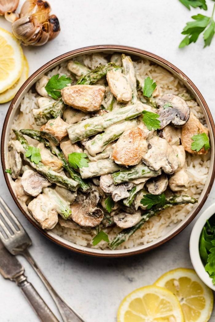 Lemon asparagus chicken in a bowl overtop rice. Next to the bowl is lemon slices, utensils, and parsley.