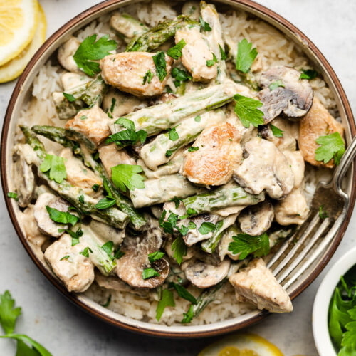Lemon garlic chicken asparagus mushroom skillet in a bowl overtop rice topped with parsley. Next to the bowl is lemon slices, a head of roasted garlic, and parsley.