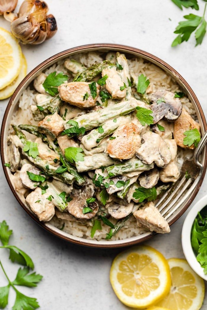 Lemon garlic chicken asparagus mushroom skillet in a bowl overtop rice topped with parsley. Next to the bowl is lemon slices, a head of roasted garlic, and parsley.