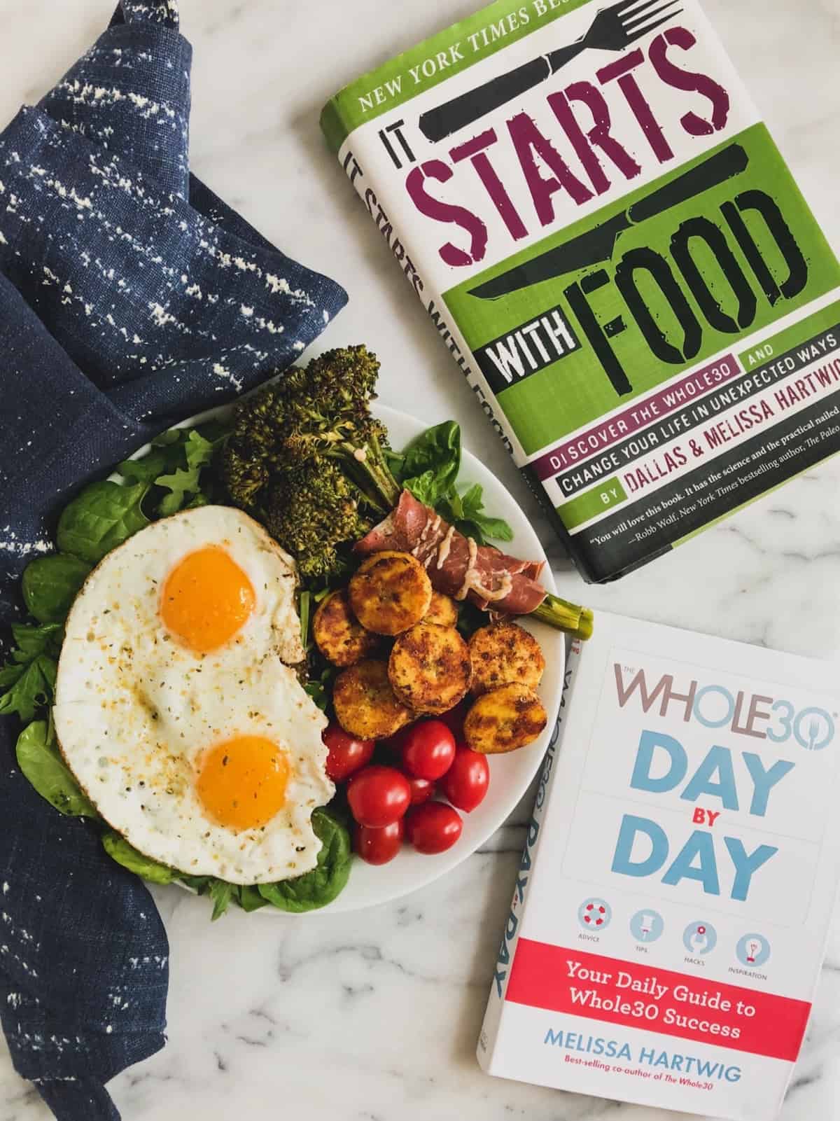 Plate with greens, fried eggs, broccolini, sautéed plantains, and cherry tomatoes next to two Whole30 books.