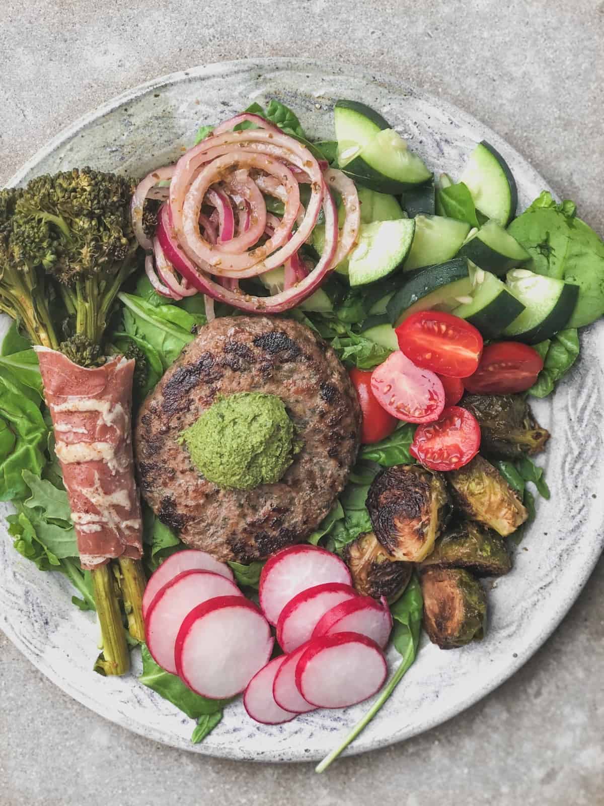 Big plate with greens, broccolini wrapped with prosuitto, marinated onions, cucumbers, tomatoes, brussels sprouts, radishes, and a burger with pesto on top