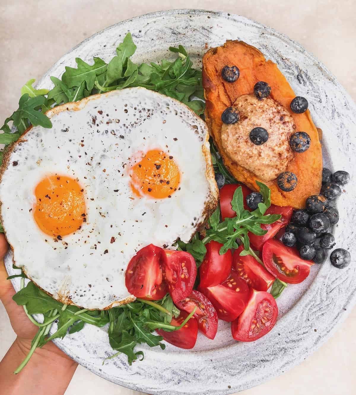 Plate with two fried eggs, half a sweet potato with almond butter and blueberries on it, and tomatoes all over greens