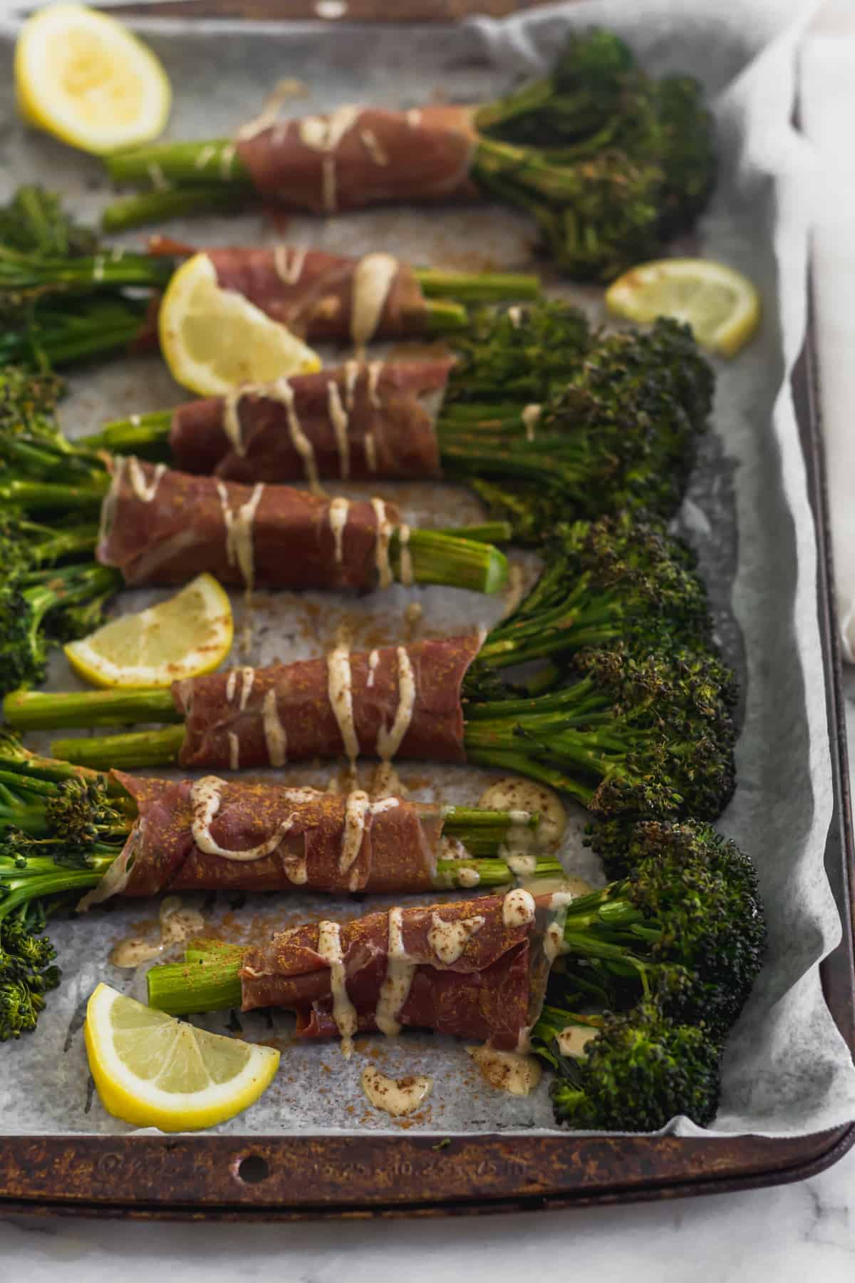 Sheet pan with a bunch of prosciutto wrapped broccolini surrounded by lemon wedges and drizzled with tahini and cinnamon