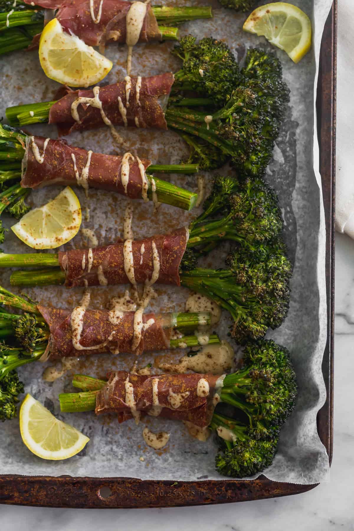 Sheet pan with a bunch of prosciutto wrapped broccolini surrounded by lemon wedges and drizzled with tahini and cinnamon