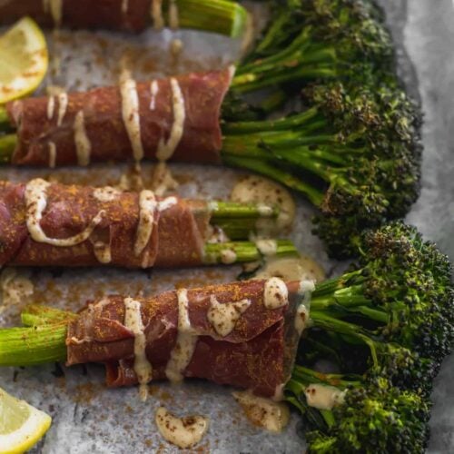 Roasted Broccolini wrapped in prosciutto on a sheet pan drizzled with tahini, lemon juice, and cinnamon