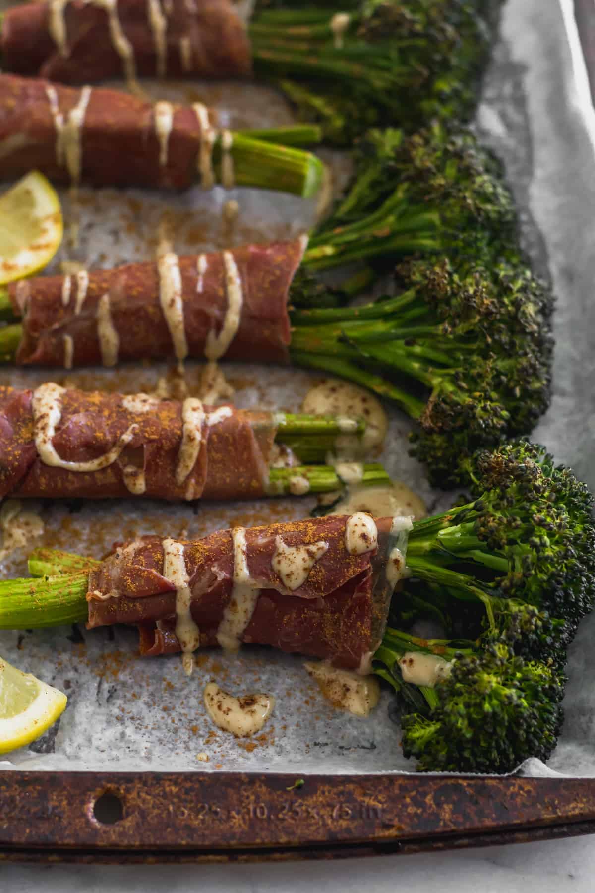 Roasted Broccolini wrapped in prosciutto on a sheet pan drizzled with tahini, lemon juice, and cinnamon
