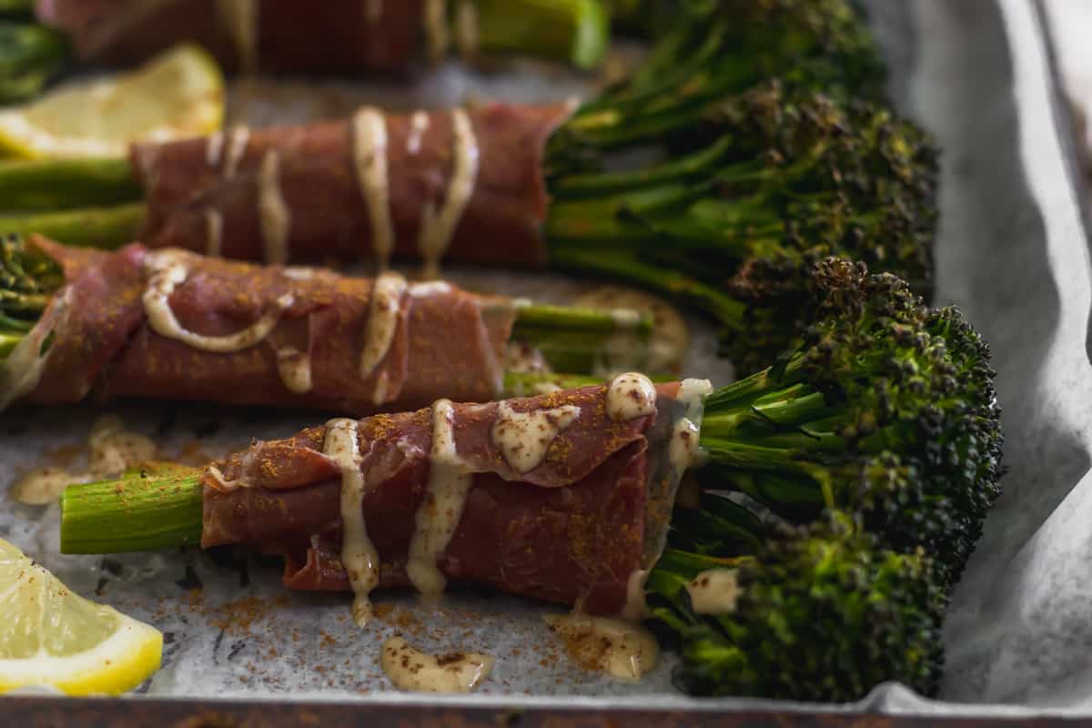 Roasted broccoli wrapped in crispy prosciutto topped with tahini and cinnamon on a sheet pan