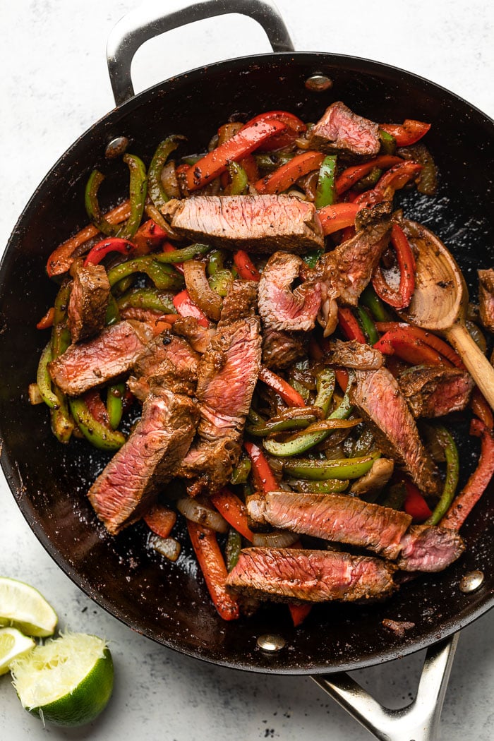 Large pan filled with cooked fajita veggies and sliced steak. Next to the pan is sliced limes.