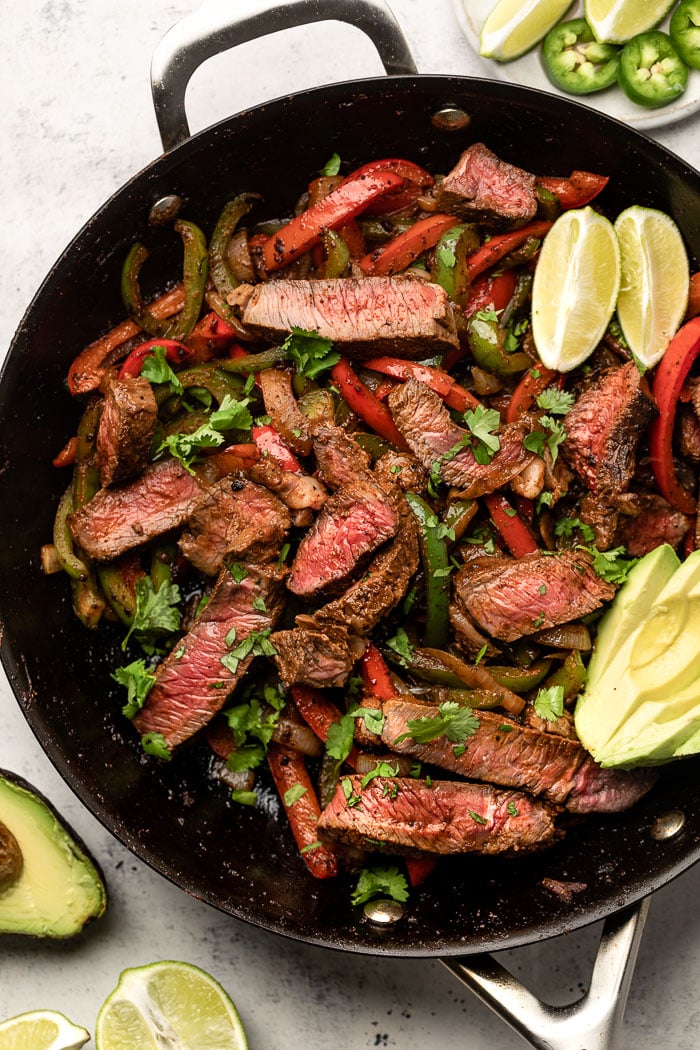 Large pan filled with healthy steak fajitas. In the pan is also some sliced avocado and lime wedges.