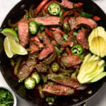 Easy steak fajitas in a large pan garnished with sliced avocado, sliced jalapeños, and lime wedges. Around it is a bowl of salsa, bowl of cilantro, and a platte with jalapeños and limes.