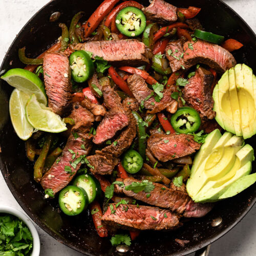 Easy steak fajitas in a large pan garnished with sliced avocado, sliced jalapeños, and lime wedges. Around it is a bowl of salsa, bowl of cilantro, and a platte with jalapeños and limes.