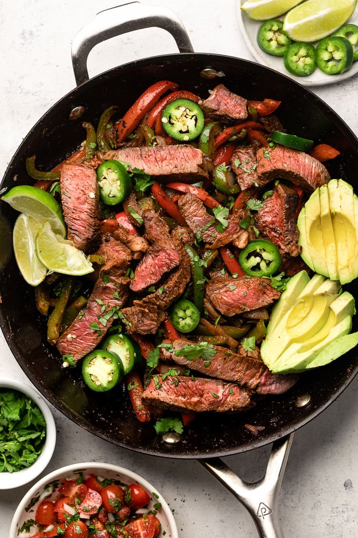 Easy steak fajitas in a large pan garnished with sliced avocado, sliced jalapeños, and lime wedges. Around it is a bowl of salsa, bowl of cilantro, and a platte with jalapeños and limes.