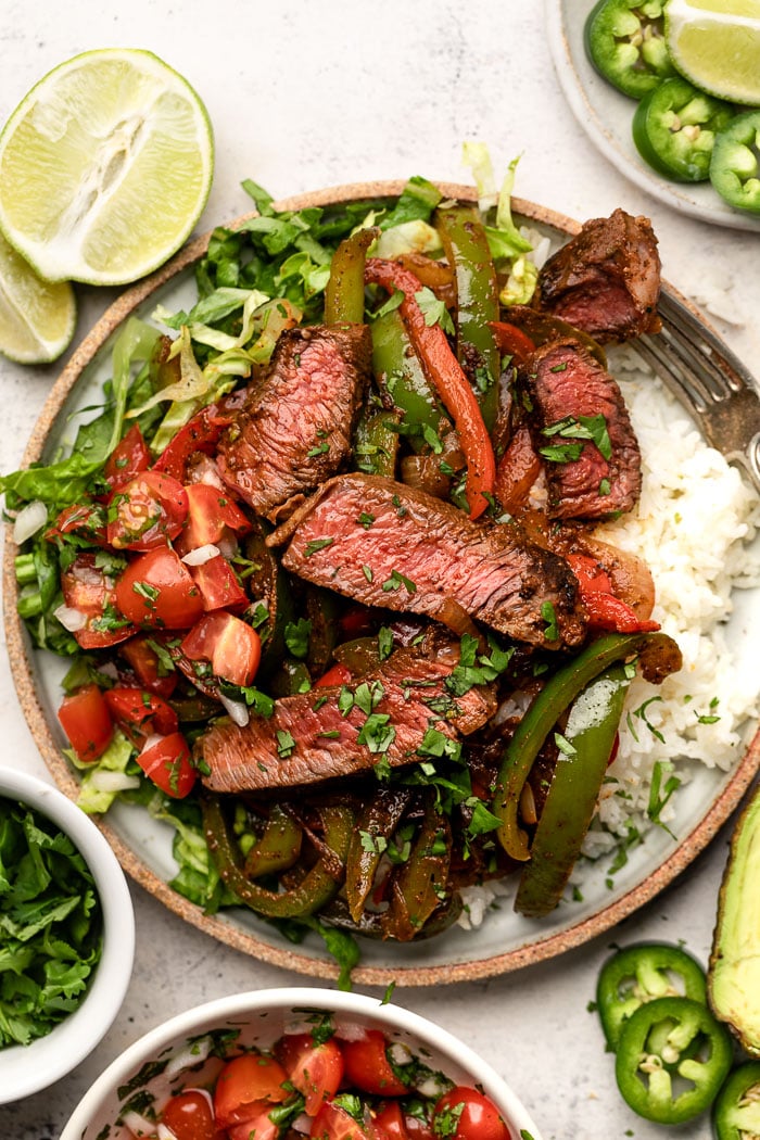 Large plate loaded with white rice, steak fajitas, chopped lettuce and tomatoes, and herbs. Around it is a bowl of diced tomatoes, bowl of cilantro, lime wedges, and a plate of sliced jalapeños.