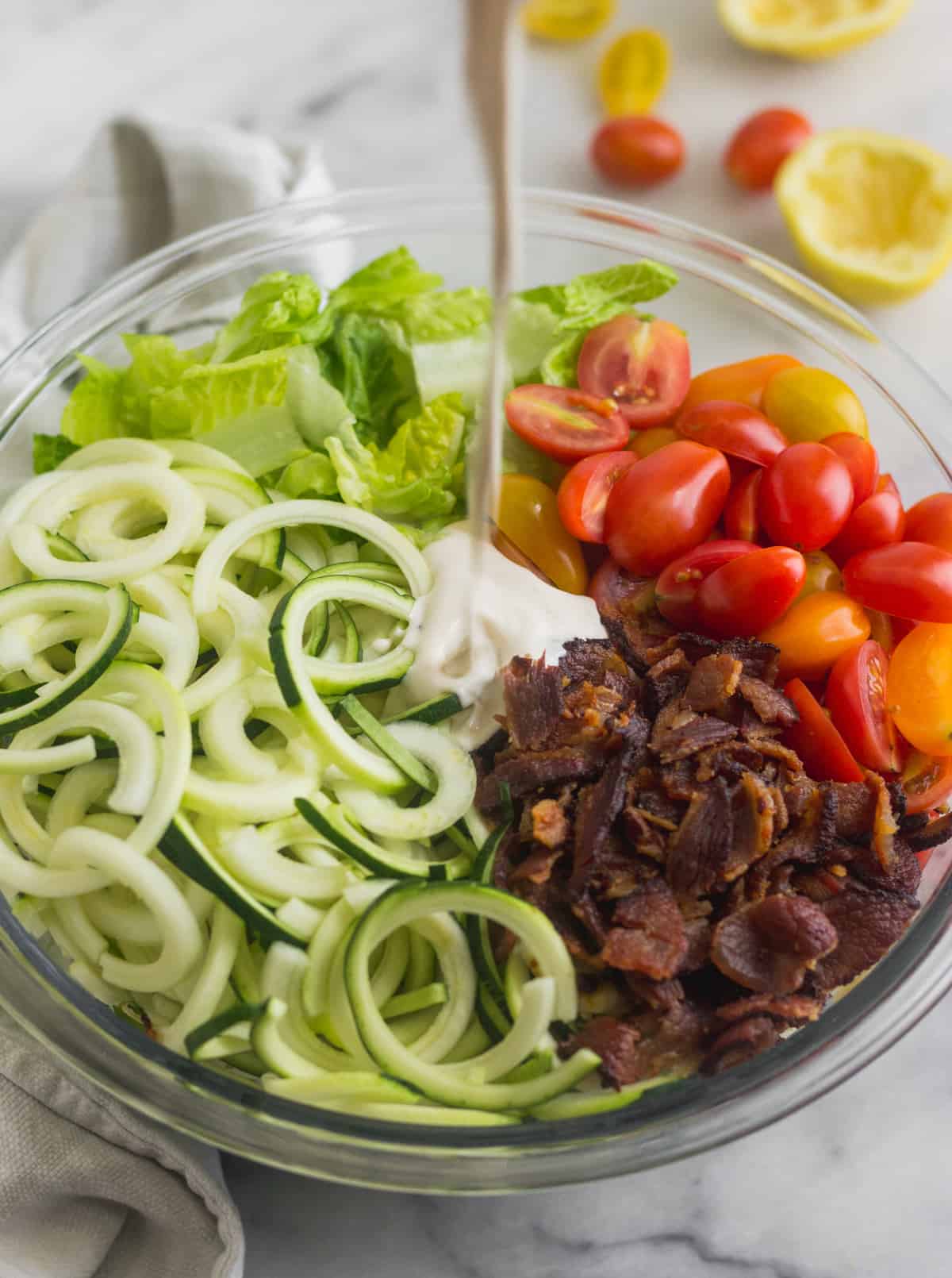 Large glass bowl with zucchini noodles, chopped romaine, cherry tomatoes, and bacon with a creamy mayo dressing being poured on it for BLT Zucchini Noodle Pasta Salad