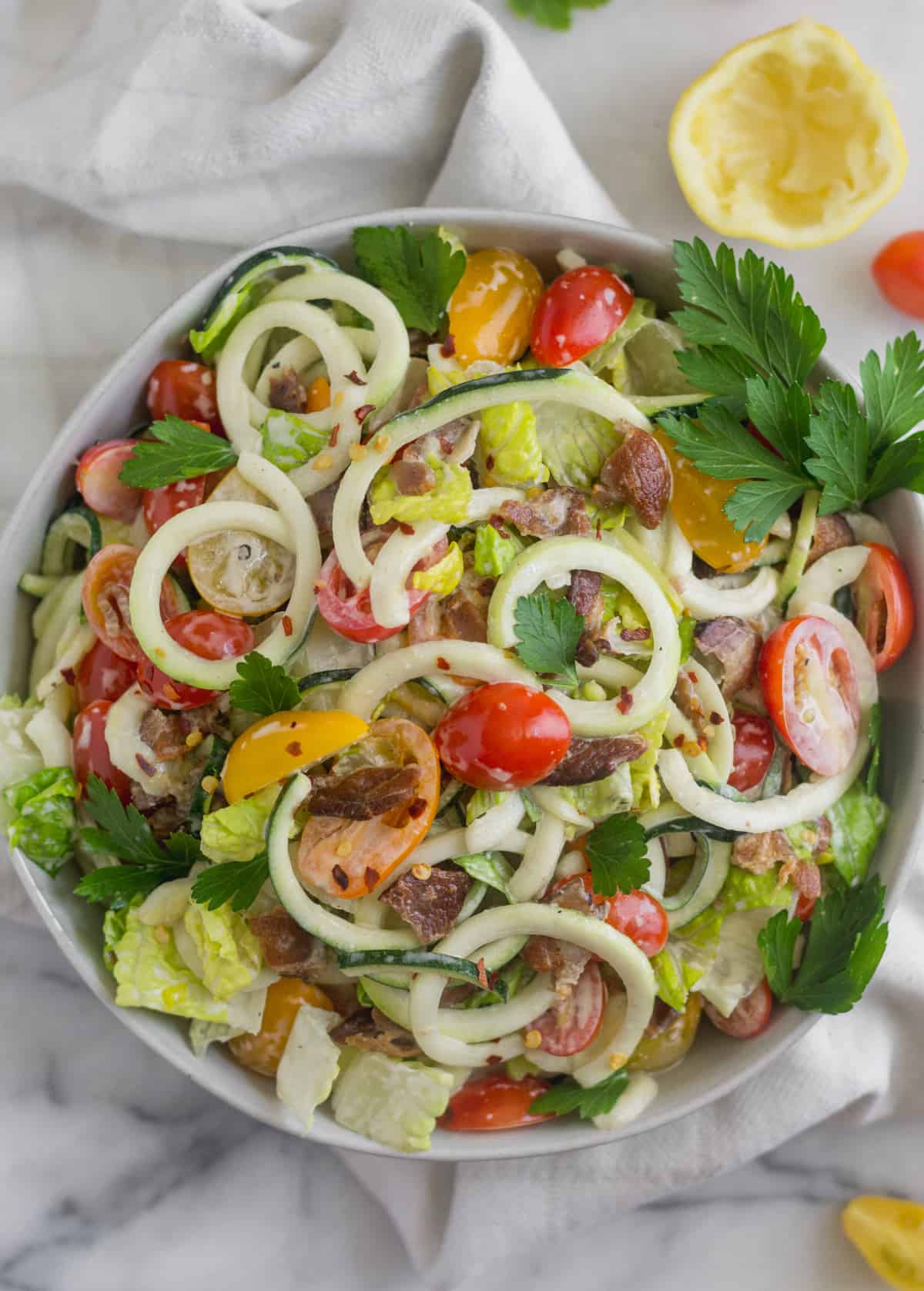 Overhead shot of Large glass bowl with zucchini noodles, chopped romaine, cherry tomatoes, and bacon for BLT Zucchini Noodle Pasta Salad in a white bowl on a white counter