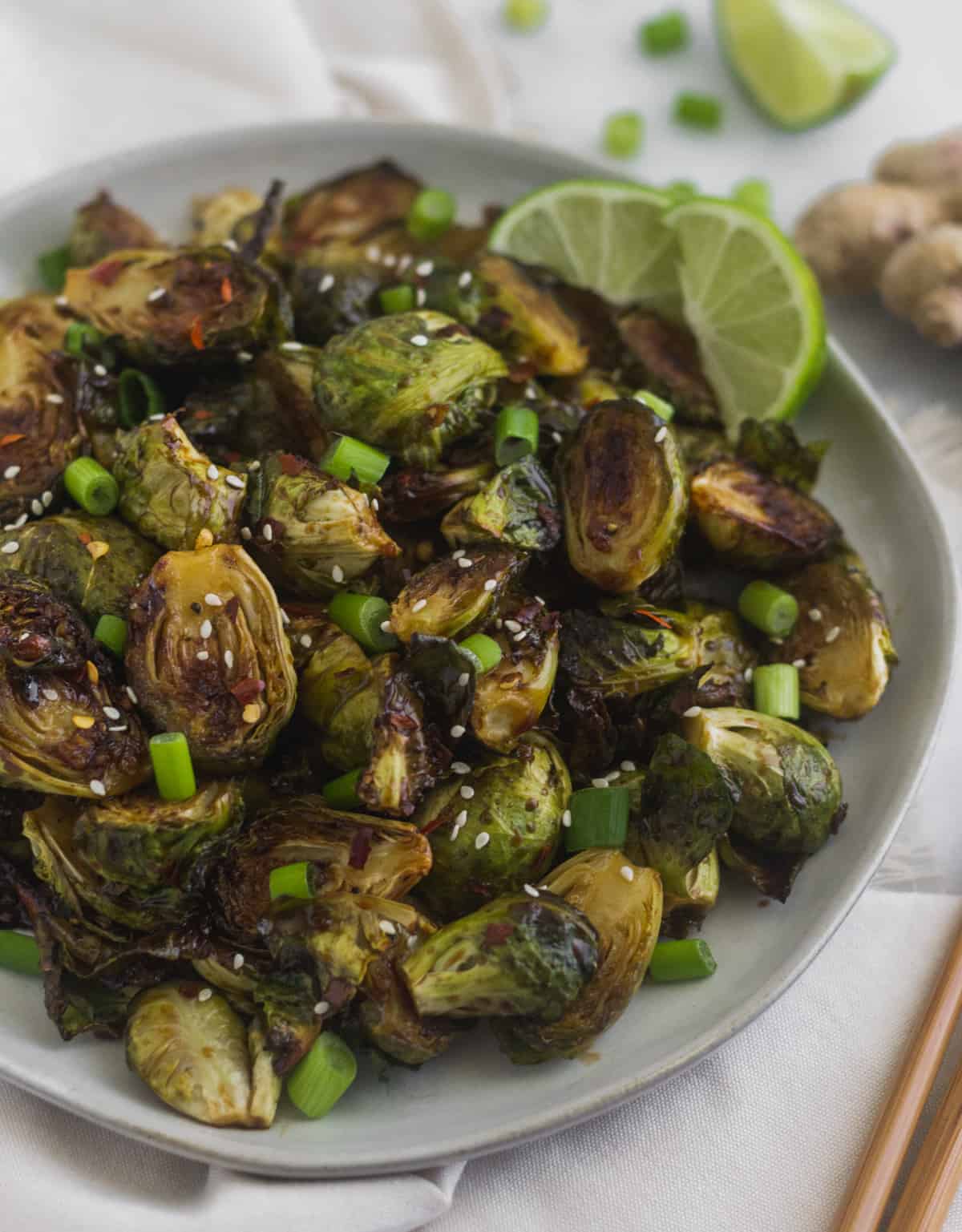 Plate of miso glazed brussels sprouts on a white linen surrounded by chopsticks, green onion, and ginger