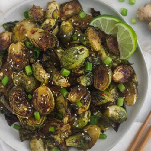 Plate of miso glazed brussels sprouts garnished with sesame seeds, green onion, and lime wedges surrounded by chopsticks and more green onions and limes