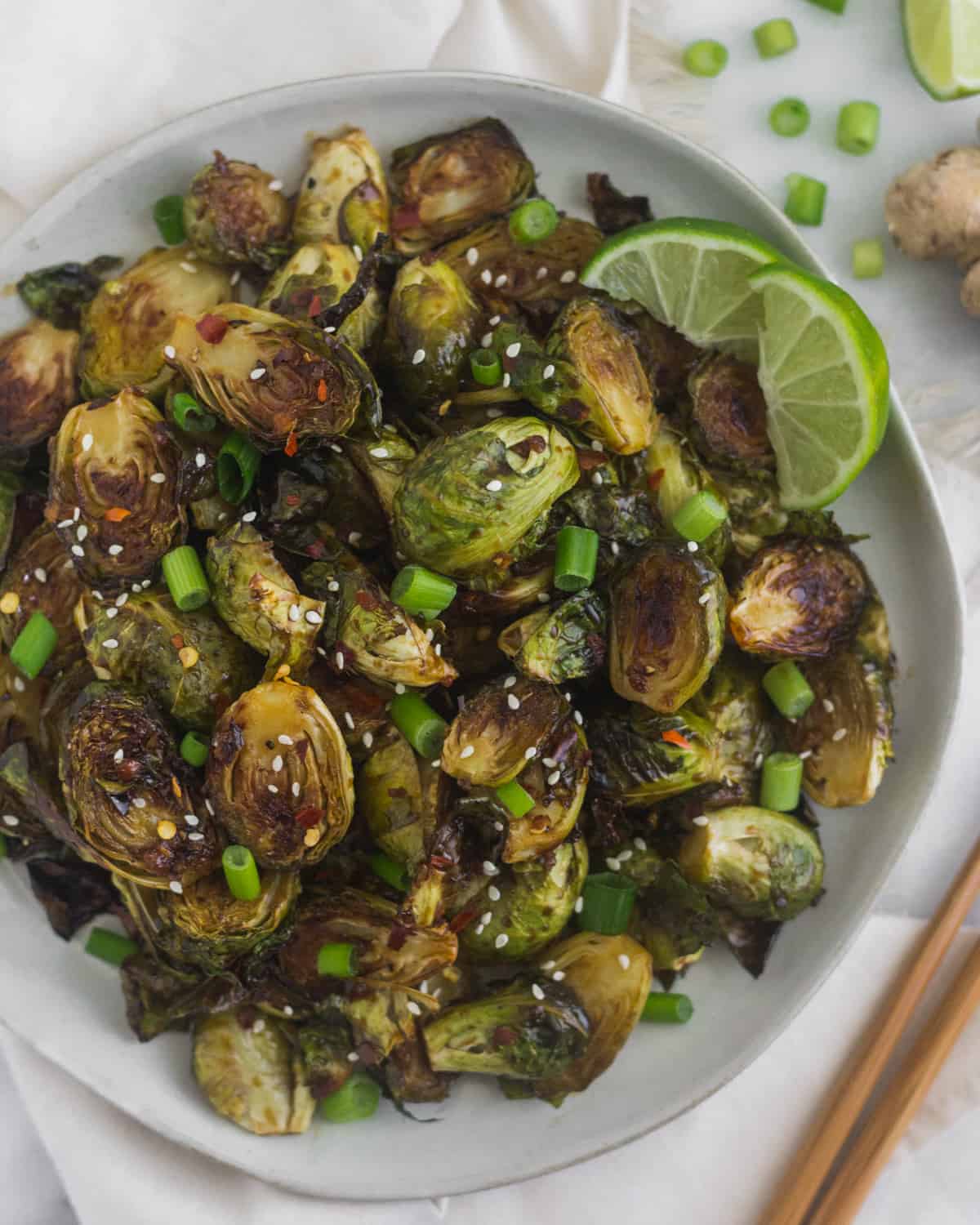 Plate of miso glazed brussels sprouts garnished with sesame seeds, green onion, and lime wedges surrounded by chopsticks and more green onions and limes