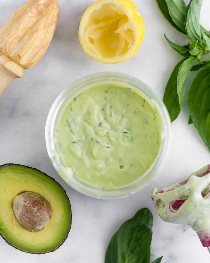 Jar filled with avocado basil aioli with an avocado, half a lemon, basil, and an immersion blender around it.