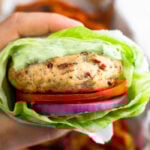 A hand holding a sun-dried tomato chicken burger wrapped with lettuce with a basket of sweet potato fries behind it.