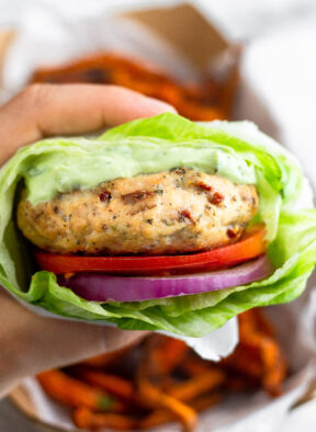A hand holding a sun-dried tomato chicken burger wrapped with lettuce with a basket of sweet potato fries behind it.
