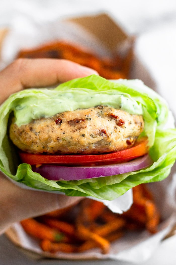 A hand holding a sun-dried tomato chicken burger wrapped with lettuce with a basket of sweet potato fries behind it.