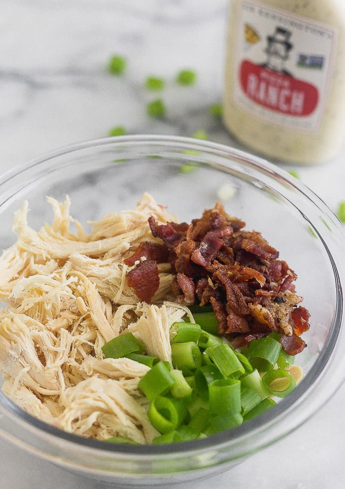 Bowl of shredded chicken, crumbled bacon, and green onions with a bottle of ranch behind it.