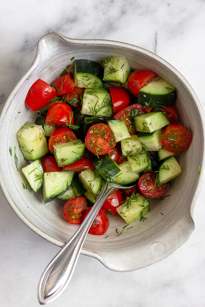 Bowl of diced cucumber and tomatoes with dill mixed in. There is a spoon sticking out of the bowl.